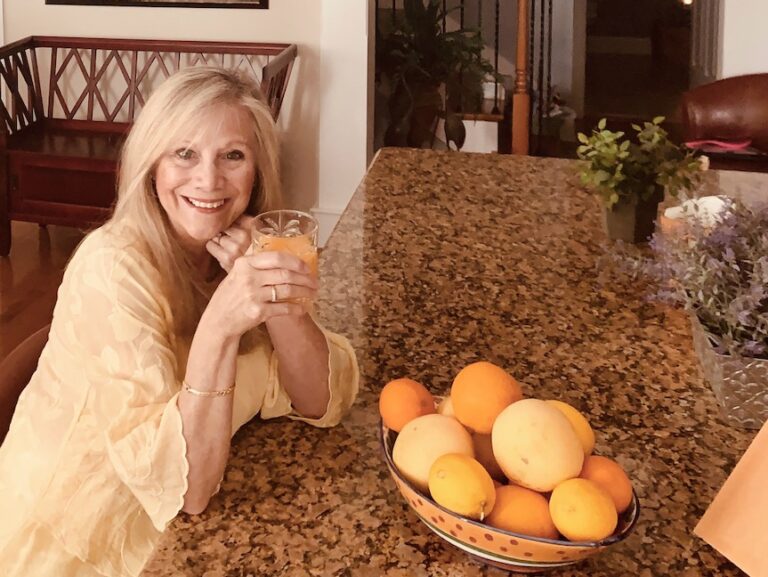 Woman sitting at the counter with a bowl of fresh citrus fruit in front of her and enjoying a glass of fresh squeezed orange juice.