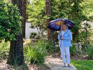 Woman standing under the trees. She is dressed for April howers wearing a blue rain slicker, rain shoes and a colorful umbrella