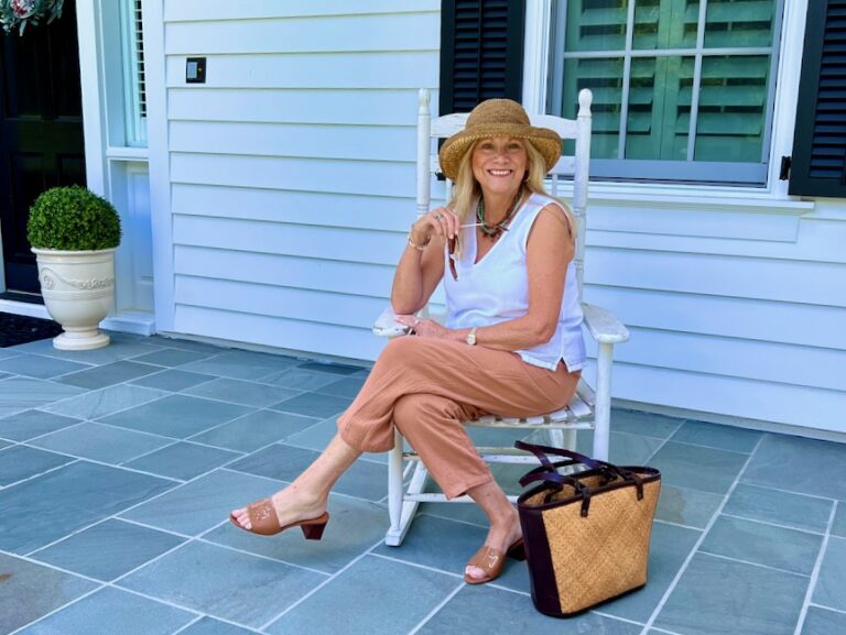 WOMAN SITTING IN A ROCKING CHAIR ON THE FRONT PORCH. SHE IS WEARING A SLEEVELESS TOP AND LINEN PANTS WITH A STRAW HAT.