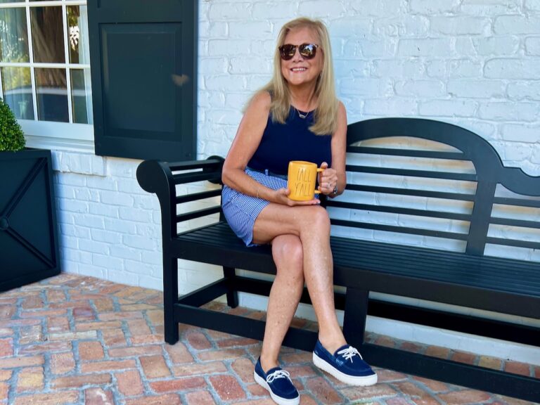 Woman sitting on a front porch bench. She is holding a coffee mug which says Fall You All