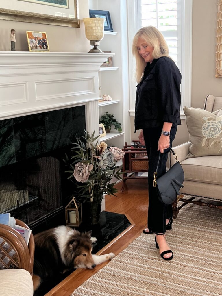A blonde woman stood before a fireplace dressed in a black jacket and denim jeans. She is looking down at a Sheltie dog.