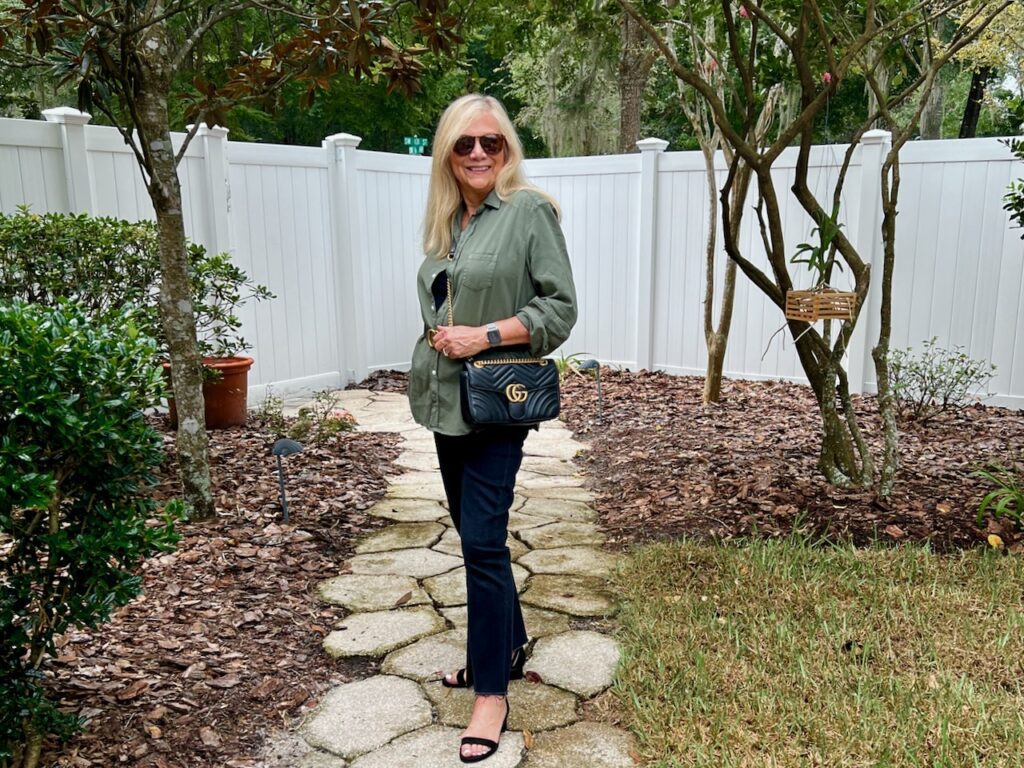 A woman standing on a garden path wearing an olive green shirt and black denim jeans.