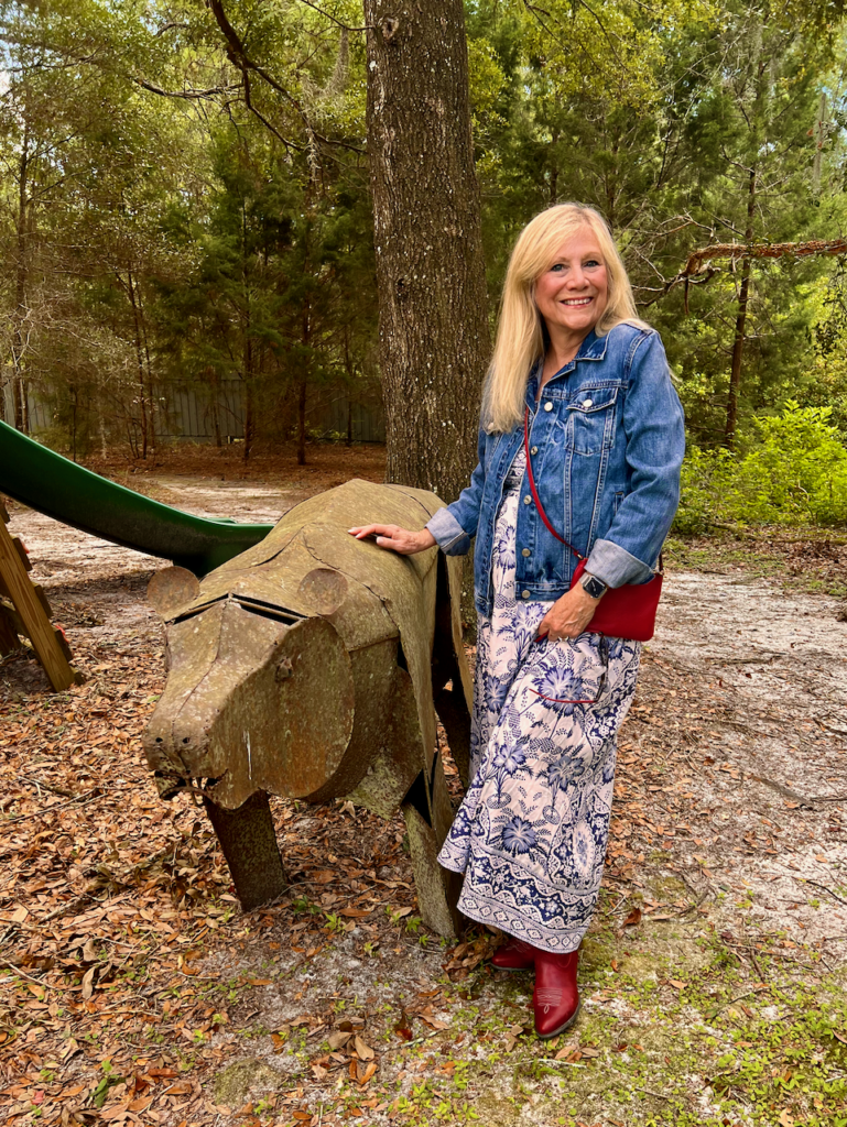 A blonde woman wearing a denim jacket is standing beside a metal sculpture of a bear. She is dressed in a style of southwestern vibes for Fall.