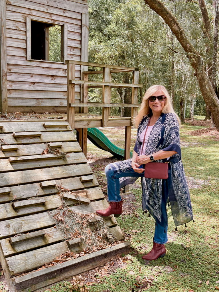 A blonde woman wearing a navy kimono, jeans, and red cowboy boots is dressed in a style of southwestern vibes for Fall. She is standing beside a wooden fort.