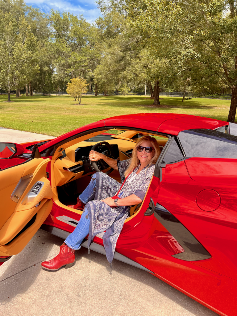 A blonde woman sitting inside a red sports car. She is wearing a southwestern fashion vibe dressed in jeans and cowgirl boots.
