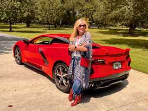 A blonde woman standing beside a red sports car dressed in a southwestern fashion vibe.