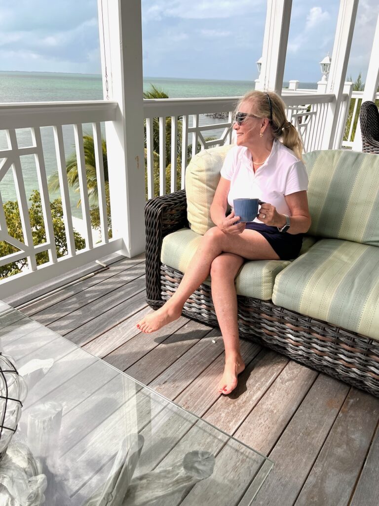 A blonde woman having coffee on the front porch and looking out at sea.