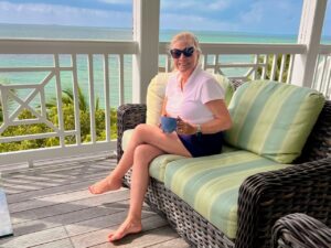 A blonde woman sitting on a front porch in a tropical ocean setting enjoying a Front Porch Coffee Chat