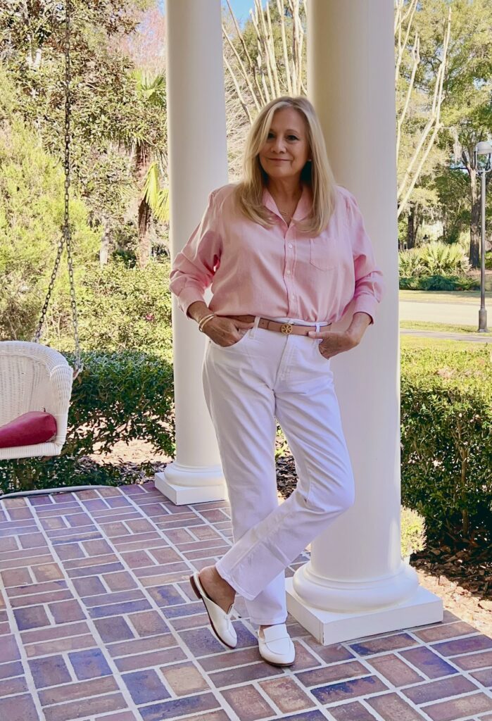 A blonde woman is standing on a front porch. She is wearing a simple style with a pink button-up shirt.