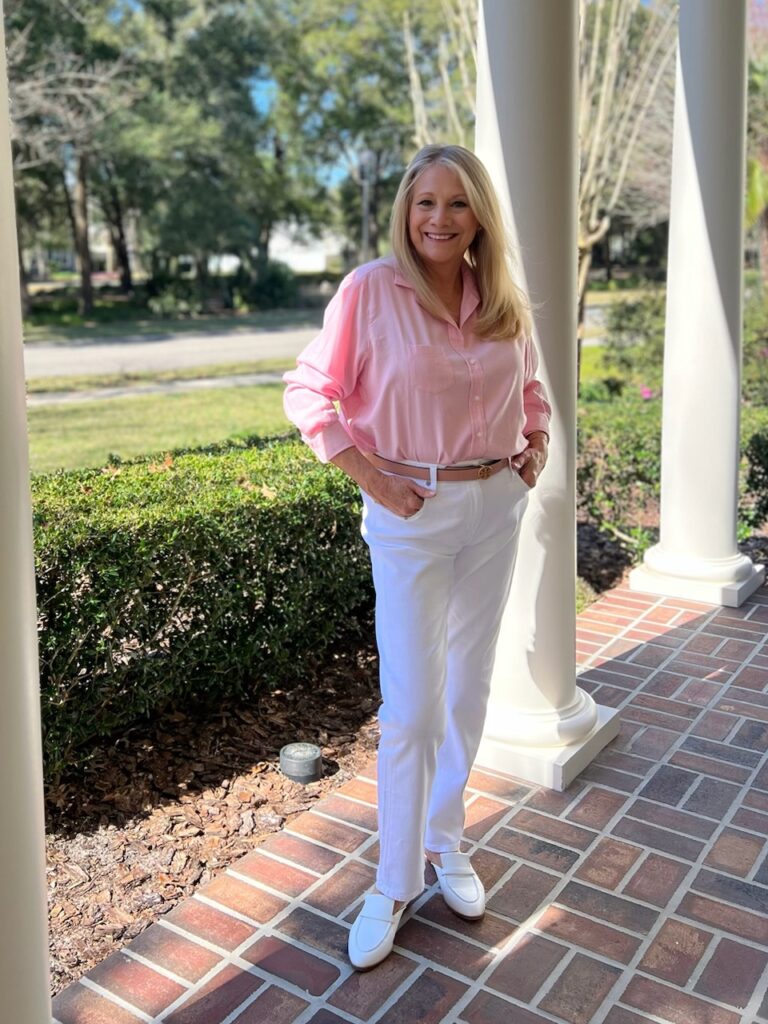 A blonde woman is standing on a front porch. She is wearing a simple style with a button-up shirt.