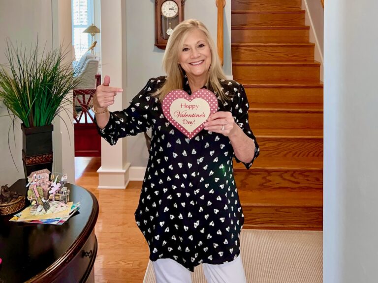 A blonde woman is wearing a black tunic shirt with white hearts. She is holding a sign, Be My Valentine,