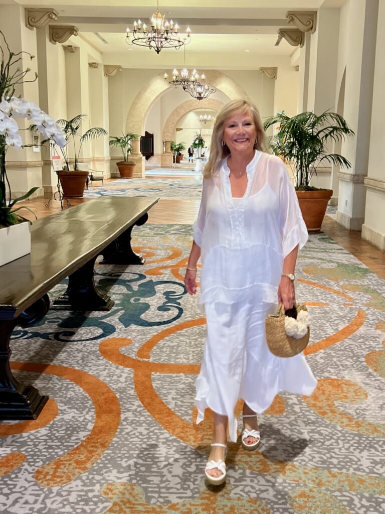 A blonde woman in a lobby is dressed in a white top and white maxi skirt.