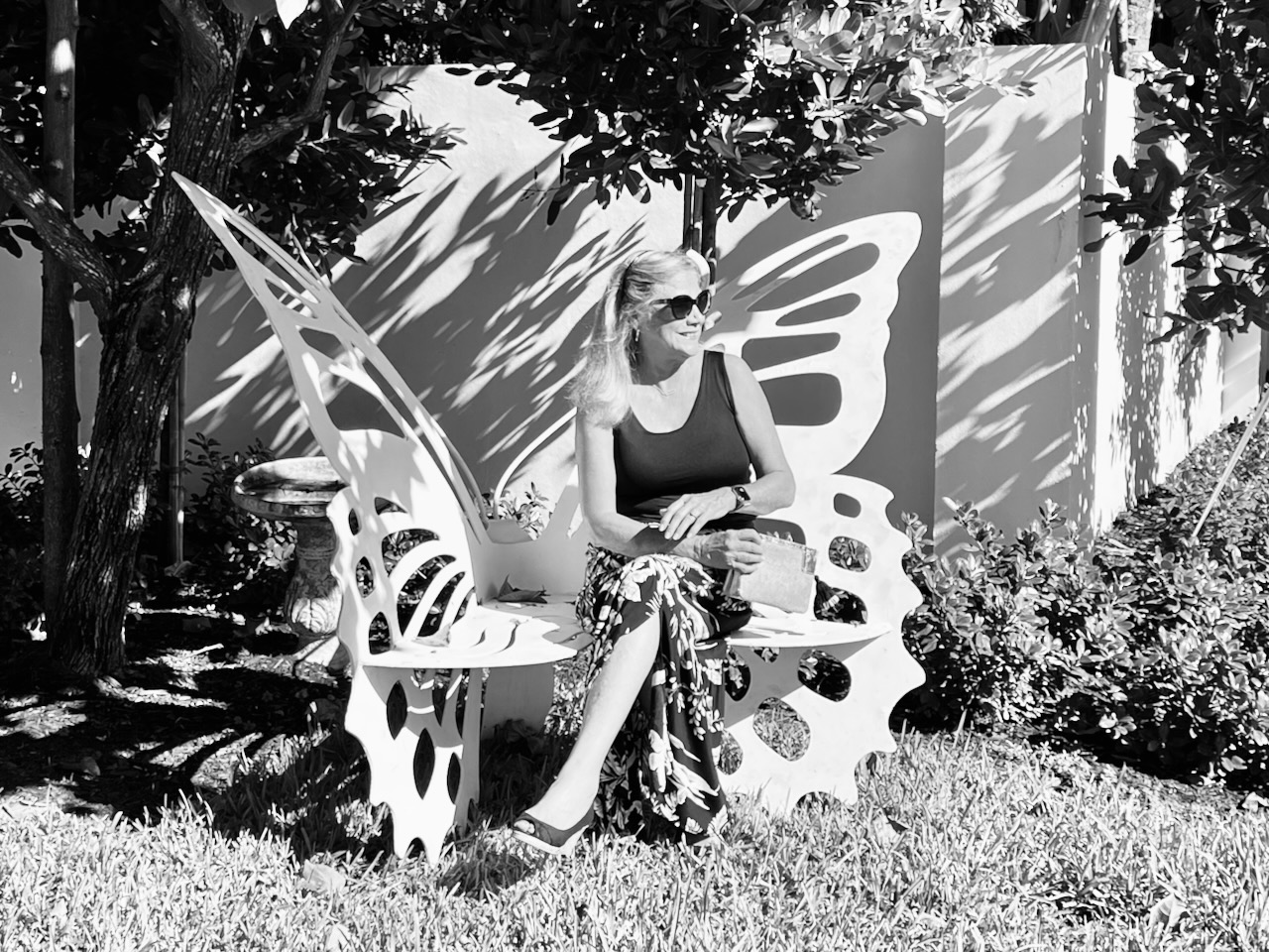 Black and white photo of a woman sitting on a butterfly bench. She is wearing a black top and a print maxi skirt.