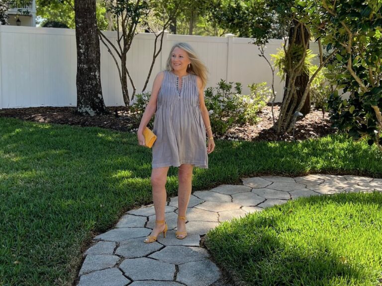 A blonde woman is walking on a stone path. She is dressed for spring, wearing yellow shoes and a handbag, as well as a gray swing dress.