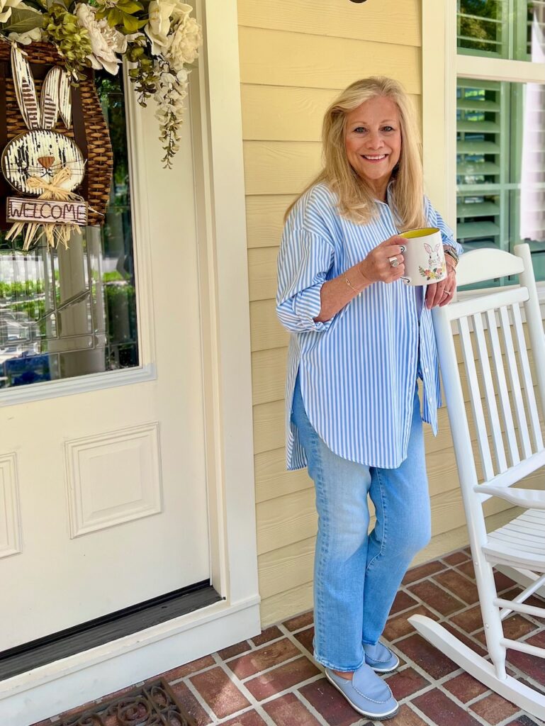 A blonde woman is standing on a porch that is decorated for Easter. She is holding a coffee cup with an Easter Bunny on the front. Front Porch Coffe Chat.