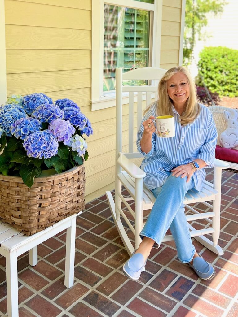 A blonde woman is sitting in a white rocking chair holding a coffee cup with an Easter Bunny on the front of it. - March Front Porch Coffee Chat.