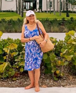 A blonde woman wearing a blue print dress, a ball cap, and holding a straw tote bag.