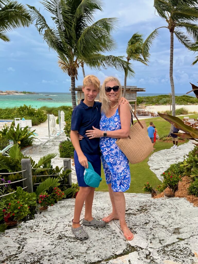 A blonde woman and a young boy are standing at a seaside resort. They are dressed in blue, and she is holding a straw tote.