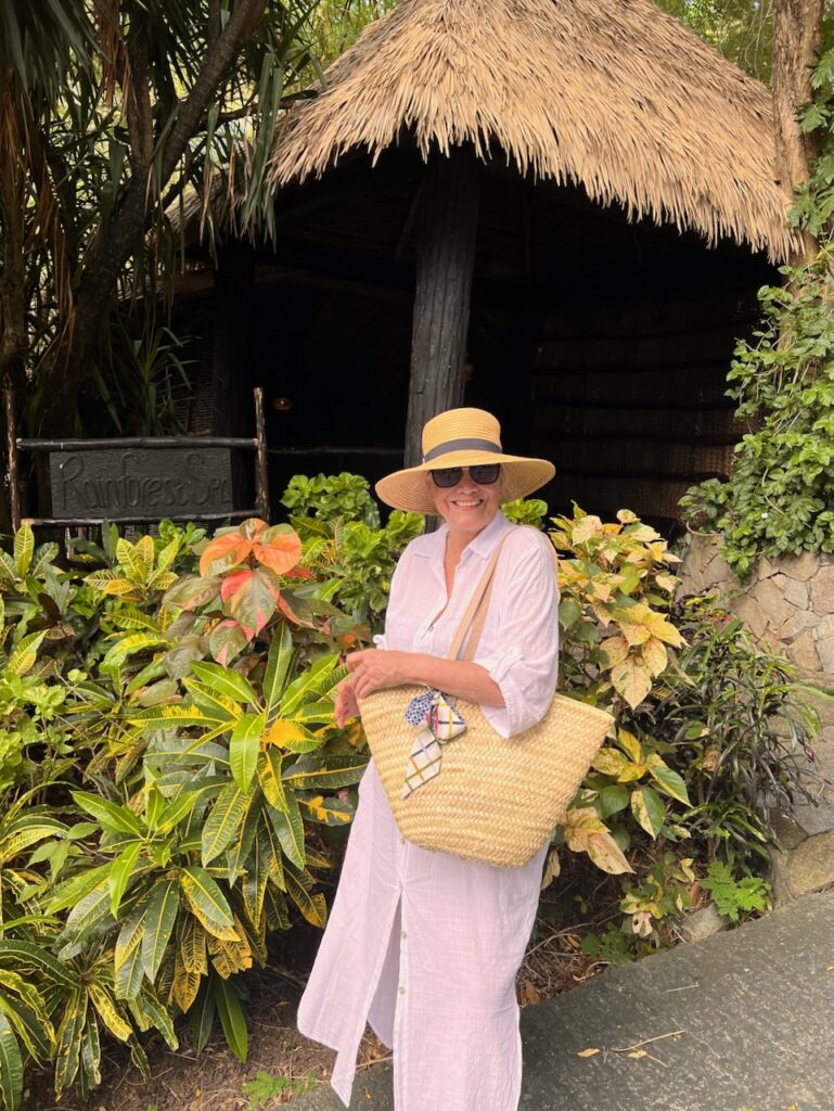 A woman in a tropical setting and she is wearing a white dress with a straw hat and straw tote.