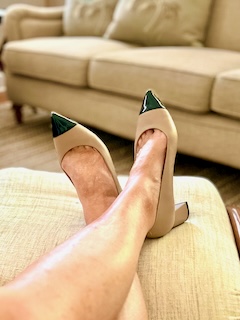 A lady's feet are on an ottoman. She is wearing Ally shoes and a pair of beige black heels.