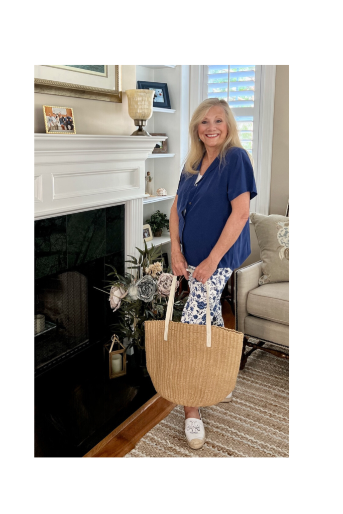 A blonde woman standing by a fireplace. She is dressed in a blue shirt with floral slacks and holding a straw tote. 