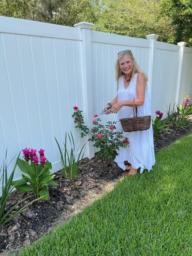 A blonde woman dressed in white with bright red sandals and accessories. She is pruning her rose bushes.