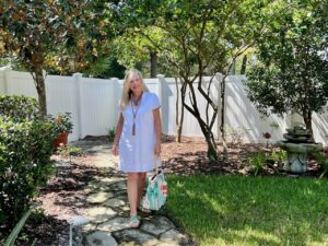 A blonde woman is walking down a stone path with a white fence in the background. She is dressed in a white dress, carrying a bright colored bag.