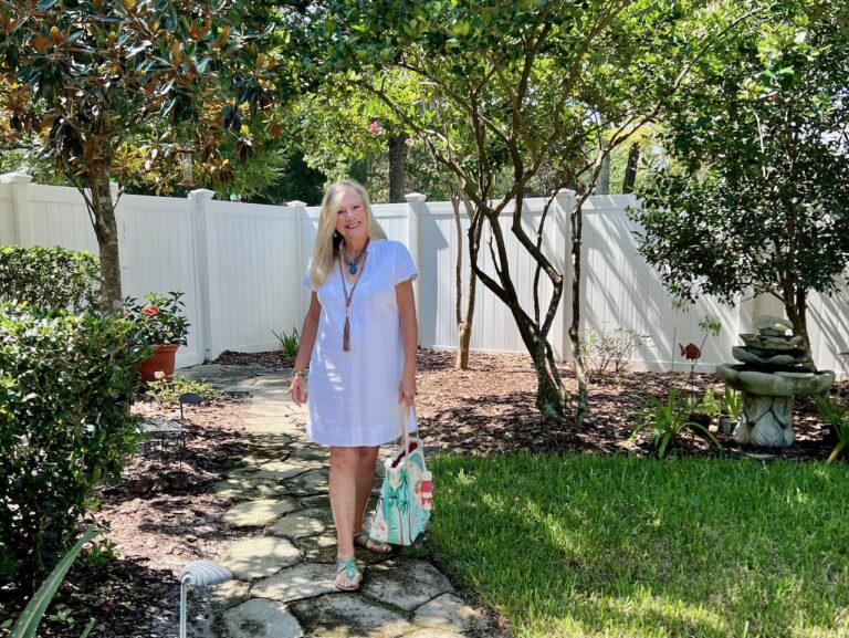 A blonde woman is walking down a stone path with a white fence in the background. She is dressed in a white dress, carrying a bright colored bag.