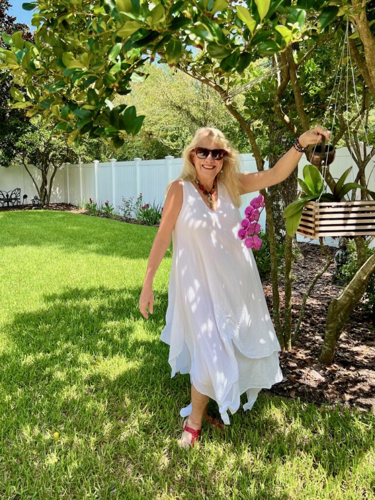 A blonde woman in a flowing white dress with bright-colored accessories is holding a watering can and watering an orchid hanging from a tree.