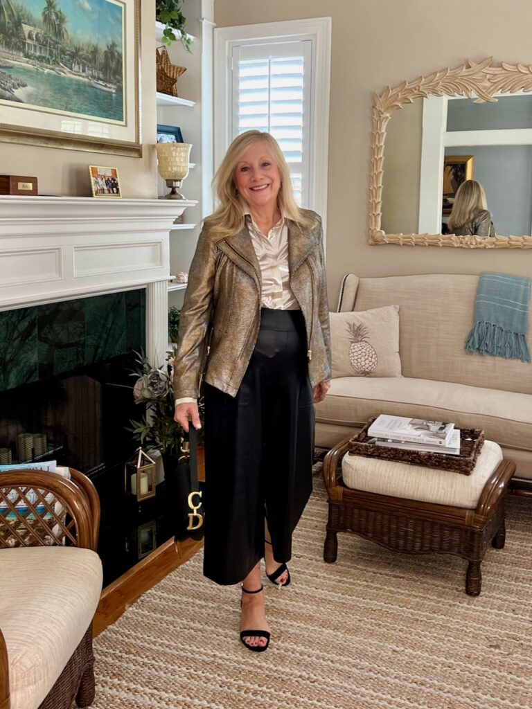 A blonde woman is standing by a fireplace. She is dressed in a Clara Sunwoo outfit of a shimmery jacket and gaucho pants.