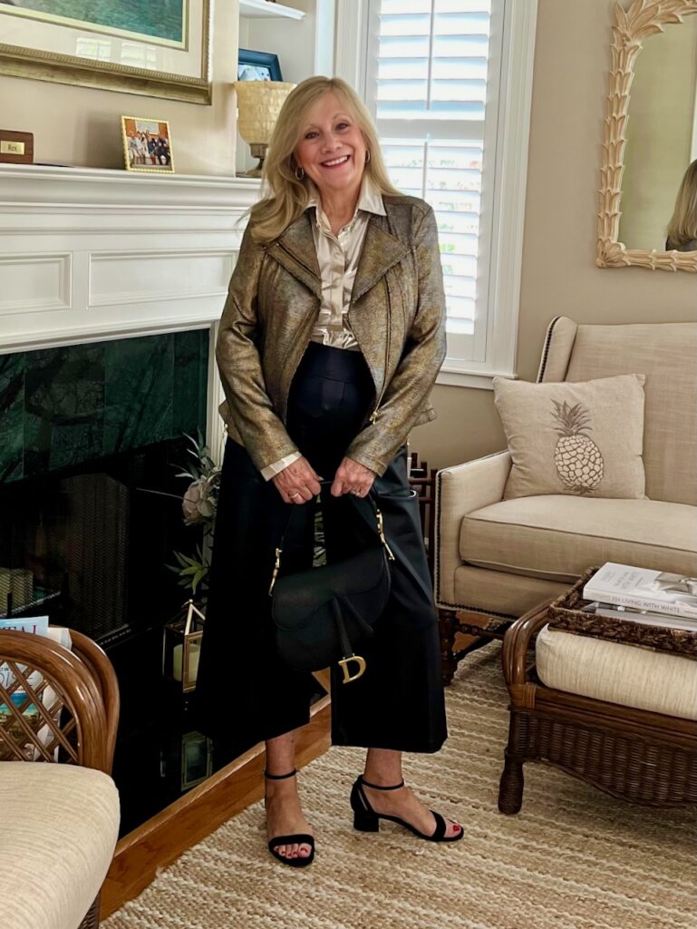 A woman wearing a leather jacket and gaucho pants while standing in front of a fireplace.