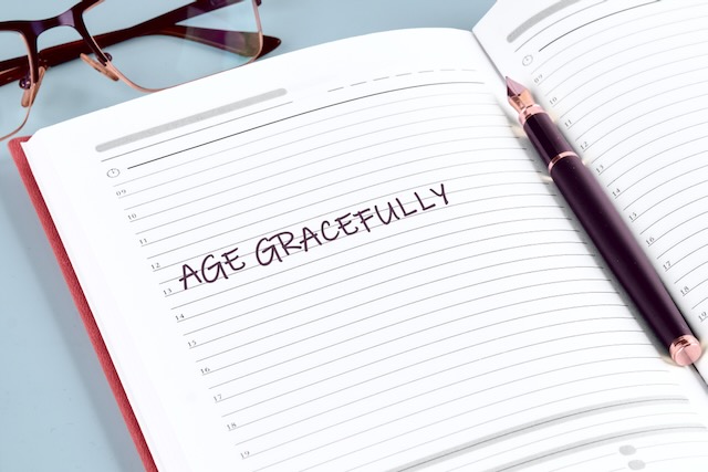 A well-organized journal lies open with the phrase age gracefully written boldly on the page. A stylish pen rests nearby, ready for notes. Glasses hint at contemplation and planning.
