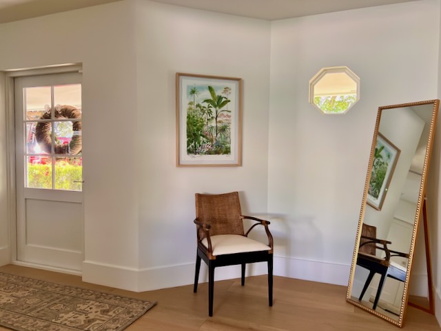 An entryway featuring a rattan chair, tropical print, and gold standing floor mirror.