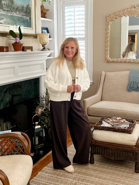 A blonde woman is standing in a living room. She is dressed in a tan top, white sweater, and brown wide-legged slacks.