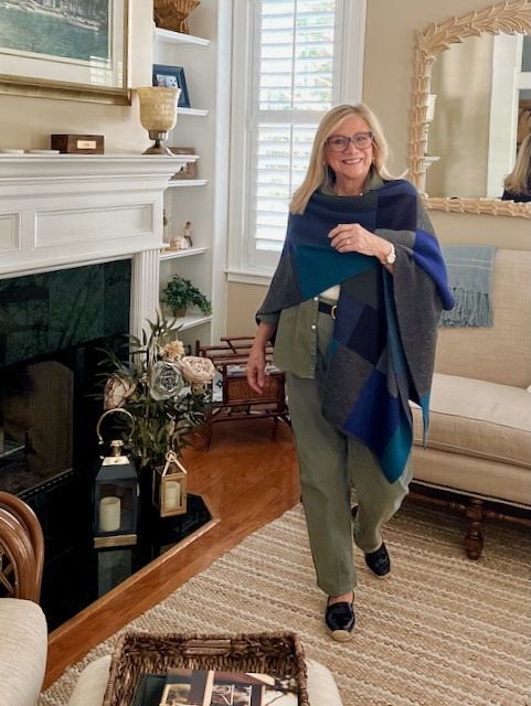 A blonde woman with glasses is wearing a wool ruana over a green shirt and pants outfit.