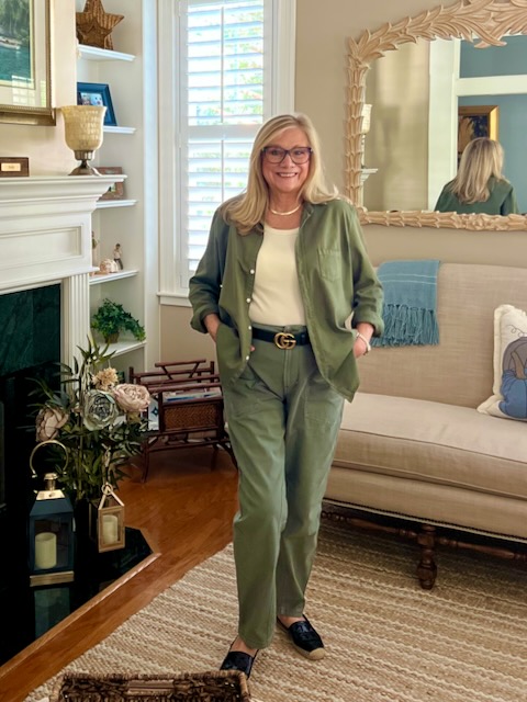 A blonde woman is standing in front of the living room fireplace. She is wearing a green shirt and pants outfit.