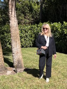 Blonde woman walking on a green lawn. She is wearing a navy blazer, sweatpants, and white top.